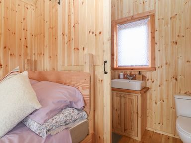 A wooden room with a bed with pillows and a sink cabinet with a window and a toilet at Hafan Gelli Sheperds Hut - Tudwal in Pwllheli