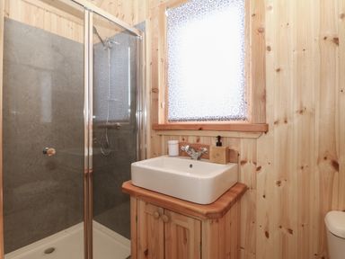 A bathroom with a shower enclosure a sink with a wooden cabinet and a window with a patterned blind at Hafan Gelli Sheperds Hut - Tudwal in Pwllheli