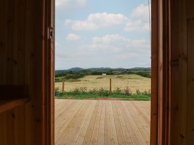 A view through an open wooden door showing a wooden deck a fenced field and hills in the distance at Hafan Gelli Sheperds Hut - Tudwal in Pwllheli