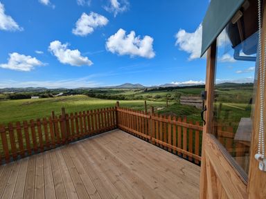 A wooden deck with wooden railing overlooking green fields and hills under a blue sky at Hafan Gelli Sheperds Hut - Tudwal in Pwllheli
