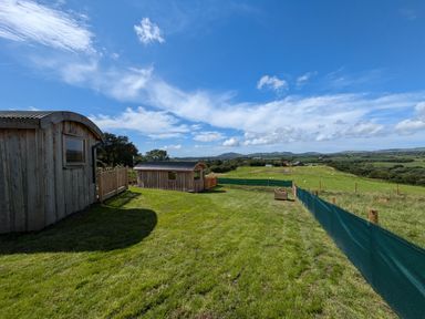 A grassy area with two wooden huts and a fenced field with hills in the background at Hafan Gelli Sheperds Hut - Tudwal in Pwllheli