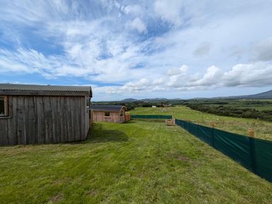 A grassy fenced yard with two wooden structures and a view of fields and hills at Hafan Gelli Sheperds Hut - Tudwal in Pwllheli