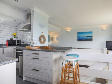 A kitchen with grey cabinets and granite countertops next to a living area with stools and a wall painting at Mor Glas in Abersoch
