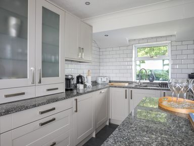 A kitchen with white cabinets gray countertops a window with a view and glassware on a tray at Mor Glas in Abersoch