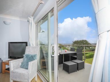 A white wicker chair with a blue cushion beside a television next to sliding glass doors opening onto a balcony with black outdoor furniture at Mor Glas in Abersoch