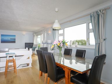 A dining area with a glass table and six chairs next to a large window with curtains and a vase of flowers at Mor Glas in Abersoch