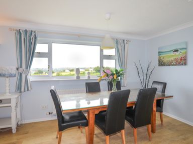 A dining room with a wooden table glass top six black chairs flowers on table window with striped curtains lamp and wall art at Mor Glas in Abersoch