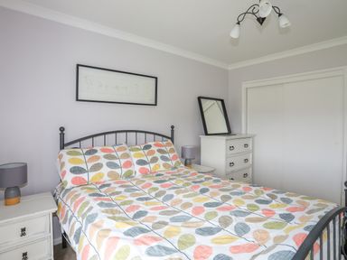 A bedroom with a bed with colorful leaf patterned bedding two bedside tables with lamps a chest of drawers with a mirror and sliding closet doors at Mor Glas in Abersoch
