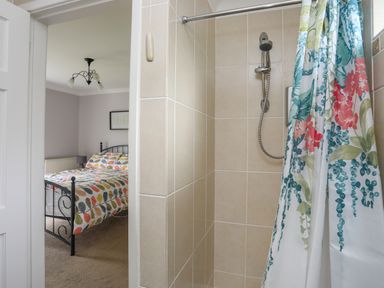 A bathroom shower with tiled walls and a floral shower curtain next to a bedroom with a bed and a lamp at Mor Glas in Abersoch