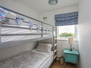 A bedroom with a bunk bed a window with a blue curtain a turquoise bedside table and a lamp at Mor Glas in Abersoch