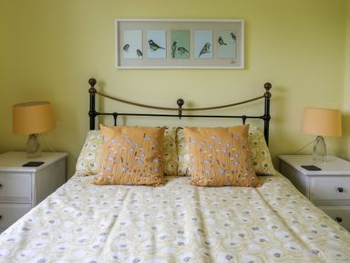 A bedroom with a bed with patterned bedding and two pillows with floral designs and two bedside tables with lamps and a framed artwork of birds on the wall at Mor Glas in Abersoch