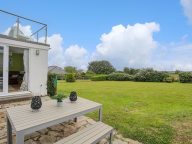An outdoor patio with a table and bench next to a grassy lawn and a house with a glass door at Mor Glas in Abersoch