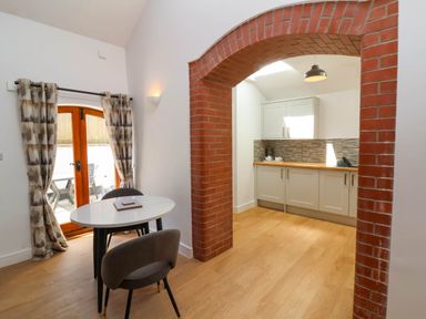 A dining area with a round table and two chairs next to a door and an open kitchen with white cabinets and wooden floor at Woodland Roost in Parkham near Bideford
