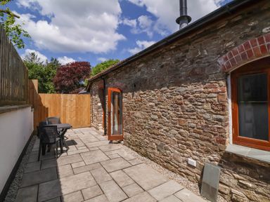 An outdoor patio area with stone paving a table with chairs a stone wall and an open wooden door at Woodland Roost in Parkham near Bideford
