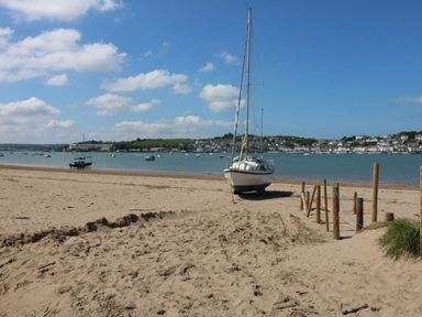 A sandy beach with a sailboat on shore and several boats in the water with buildings in the background at Woodland Roost Parkham near Bideford
