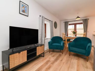 A living room with a television on a wooden cabinet two blue armchairs a wooden dining table with chairs and flowers at Ladybird Rest in Parkham near Bideford