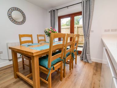 A dining room with a wooden table and six chairs with cushions and placemats with glass doors and curtains at Ladybird Rest in Parkham near Bideford