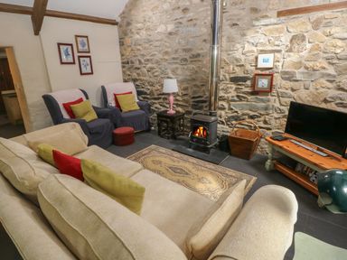 A living room with a stone wall and wood burning stove with beige sofa armchairs and a television at The Old Stable in Pontfaen near Fishguard