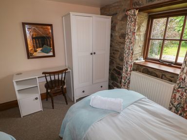 A bedroom with a bed a towel a desk a wooden chair a wardrobe and a window with floral curtains at The Old Stable in Pontfaen near Fishguard