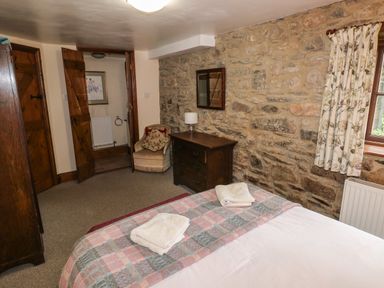 A bedroom with a bed, wooden chest of drawers, armchair, stone wall, window with floral curtains and wooden door at The Old Stable Pontfaen near Fishguard
