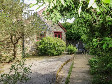 A stone house with red framed windows and doors surrounded by green bushes and trees with a patio and outdoor chairs at The Old Stable in Pontfaen near Fishguard