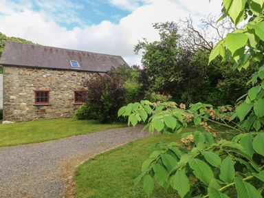 A stone building with red window frames near a gravel path and green trees at The Old Stable in Pontfaen near Fishguard