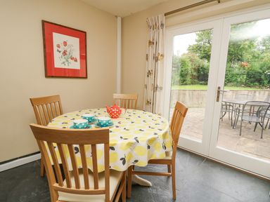 A dining area with a round table covered with a yellow patterned tablecloth surrounded by four wooden chairs next to glass doors leading to a patio at Beachway Brynhenllan near Dinas Cross