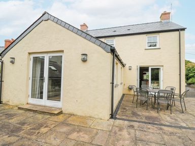 An outdoor patio area with a metal table and six chairs outside a beige house at Beachway Brynhenllan near Dinas Cross