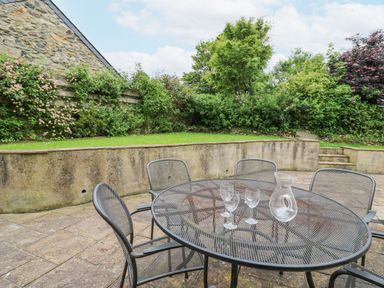 An outdoor patio with a round metal table and chairs three glasses and a glass pitcher on the table a stone wall plants and trees at Beachway Brynhenllan near Dinas Cross