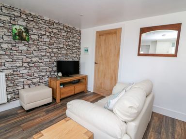 A living room with a stone patterned wallpaper wall a wood tv stand with a television a beige ottoman a cream sofa and a mirror on the wall at Field View in Llanybydder