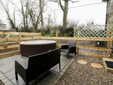 An outdoor patio area with a hot tub two black wicker chairs a wooden fence and gravel at Field View in Llanybydder
