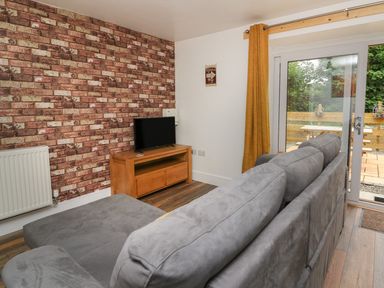 A living room with a grey sofa a wooden TV stand with a TV brick pattern wallpaper and sliding glass door at Garden View in Llanybydder