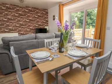 A dining table set for four with plates glasses and a vase of flowers next to a grey sofa and sliding glass doors at Garden View in Llanybydder