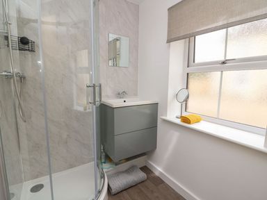 A bathroom with a curved glass shower enclosure a gray wall-mounted sink cabinet a small mirror and a window with a yellow towel on the sill at Garden View in Llanybydder