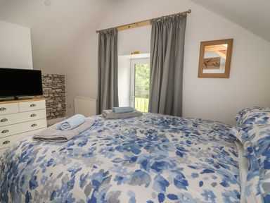 A bedroom with a bed covered in blue and white floral bedding towels on the bed a dresser with a TV a framed picture on the wall and a window with gray curtains at Garden View in Llanybydder