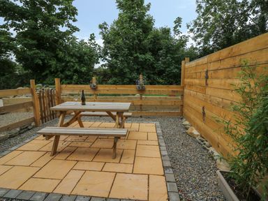 An outdoor patio area with a wooden picnic table with a bottle and glass on it surrounded by wooden fences and trees at Garden View in Llanybydder
