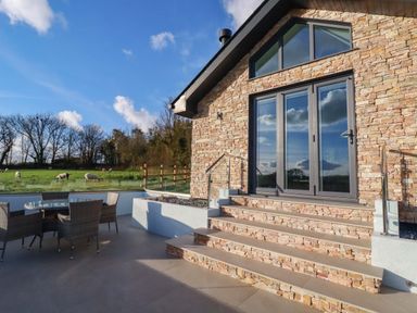 An outdoor patio area with a stone house and glass doors next to a table with chairs and a grassy field with sheep at Lake View in Kestle Mill near Quintrell Downs