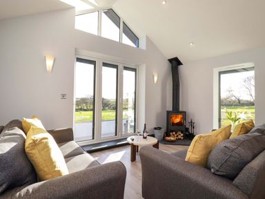 A living room with gray sofas and yellow pillows a round wooden table a wood burning stove and large windows at Lake View in Kestle Mill near Quintrell Downs