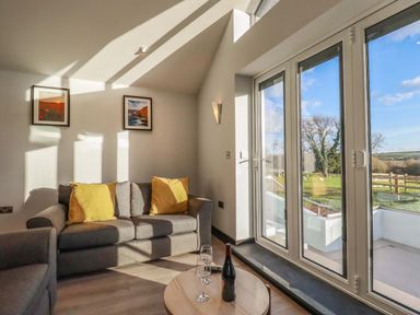 A living room with a gray sofa and yellow cushions and a round wooden table with a wine bottle and glasses near large glass doors at Lake View in Kestle Mill near Quintrell Downs