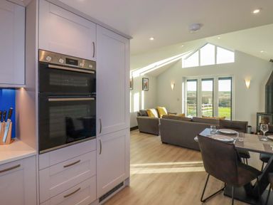 A kitchen with an oven unit and knife block next to a dining table and chairs in an open living space with sofas and large windows at Lake View in Kestle Mill near Quintrell Downs