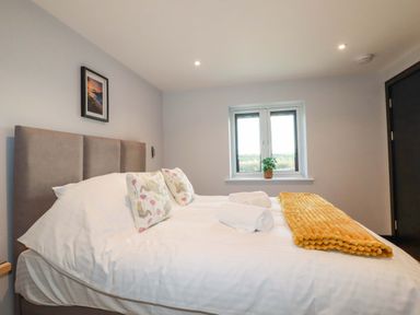 A bedroom with a double bed pillows a rolled towel and a yellow blanket on the bed a window with a plant on the sill and a framed picture on the wall at Lake View in Kestle Mill near Quintrell Downs