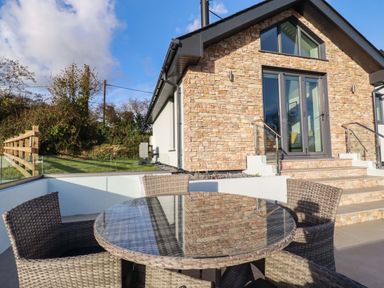 An outdoor patio with a glass top round table and four wicker chairs next to a stone wall house at Lake View in Kestle Mill near Quintrell Downs