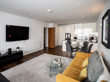 An open living room and kitchen with a yellow sofa, glass coffee table, dining table with chairs, and wall-mounted TV at Apartment 17 Abbey Sands in Torquay