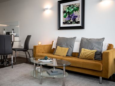 A living room with a mustard sofa gray and yellow cushions glass coffee table and dining chairs at Apartment 17 Abbey Sands Torquay