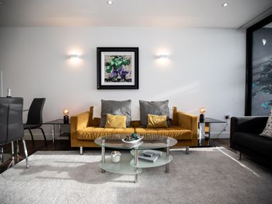 A living room with a mustard sofa gray cushions glass coffee table and black dining chairs at Apartment 17 Abbey Sands in Torquay