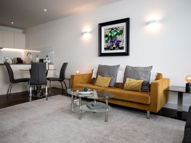 A living room with a mustard sofa gray cushions and a glass coffee table next to a kitchen with black dining chairs at Apartment 17 Abbey Sands in Torquay