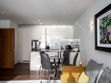 A kitchen with white cabinets and a glass dining table with four chairs next to a yellow sofa with cushions at Apartment 17 Abbey Sands in Torquay