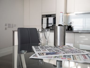 A kitchen with a glass table holding a bottle in an ice bucket two empty glasses and newspapers at Apartment 17 Abbey Sands in Torquay