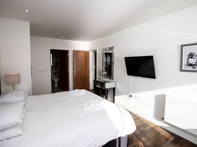 A bedroom with a bed, bedside lamp, wall-mounted TV, dressing table, and open doors showing a bathroom at Apartment 17 Abbey Sands in Torquay