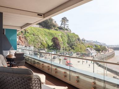 A balcony with chairs and a small grill overlooking a promenade and hillside at Apartment 17 Abbey Sands in Torquay
